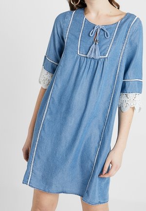 Denimmekko, jossa on pyöreä pääntie, jossa on tupsuja, lyhyet hihat valkoisilla pitsikoristeilla ja pystysuorat ompeleet sivuilla.