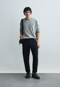 Grå t-shirt med rund hals och korta ärmar, kombinerad med svarta plisserade byxor. Accessoarerna består av en svart axelväska och svarta sneakers.