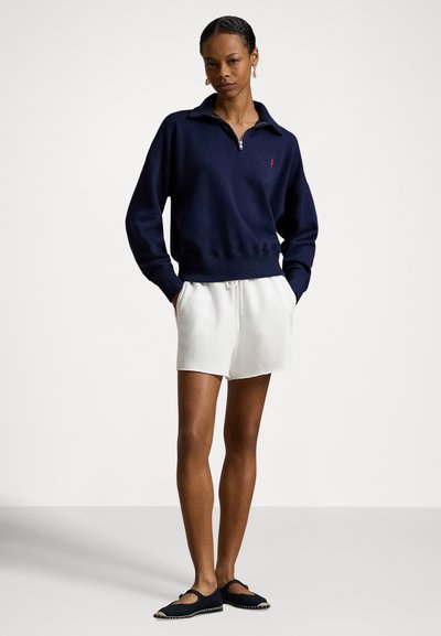 Γυναίκα όρθια με τα χέρια στις τσέπες, φοράει μπλε ζακέτα με φερμουάρ, λευκά σορτς με κορδόνι και μαύρες παντόφλες, μπροστά σε απλό φόντο.