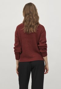 Nainen, jolla on aaltoileva vaaleanruskea hius, pukeutuneena teksturoituun tummanpunaiseen neuleeseen ja mustiin housuihin, seisoo yksinkertaista vaaleanharmaata taustaa vasten.