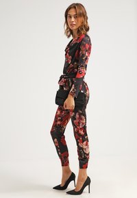 Jumpsuit floreale in nero con motivi rossi e rosa, con vita cinta e maniche lunghe. Abbinato a tacchi neri e una pochette.