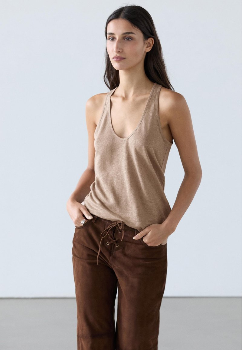 Woman with long dark hair wearing a beige sleeveless V-neck top and brown lace-up pants, standing with hands in pockets against a plain background.