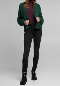 Cardigan verde texturizado sobre un suéter de tortuga color marrón, combinado con jeans ajustados negros y botas negras hasta el tobillo con suela gruesa.