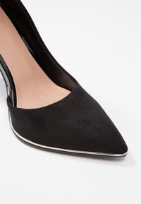 Decolleté in suede nero a punta con un pannello laterale lucido nero e rifiniture argentate lungo la suola. Fodera interna morbida.