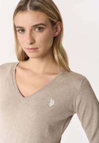 Maglione beige a scollo a V con collo a coste e un piccolo dettaglio logo bianco sul lato sinistro del petto. Realizzato in un morbido materiale knit leggero.