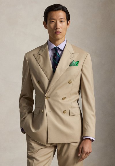 Un uomo indossa un abito doppiopetto beige con un fazzoletto da taschino verde decorato, cravatta a righe e una camicia viola chiaro, in piedi con una mano in tasca.