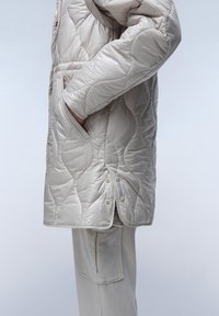 Cappotto imbottito trapuntato beige chiaro con colletto alto, tasche laterali, chiusure a scatto e finitura texturizzata. Abbinato a pantaloni chiari coordinati.