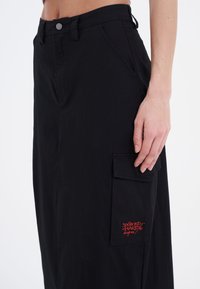 Sort cargoskirt med høj talje, sidelommer og et lille rødt broderet logo på sidelommen. Glat stof og afslappet pasform.