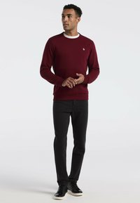 Pull bordeaux à manches longues avec un col rond, associé à un pantalon noir slim et des chaussures noires. Présente un petit logo sur la poitrine.