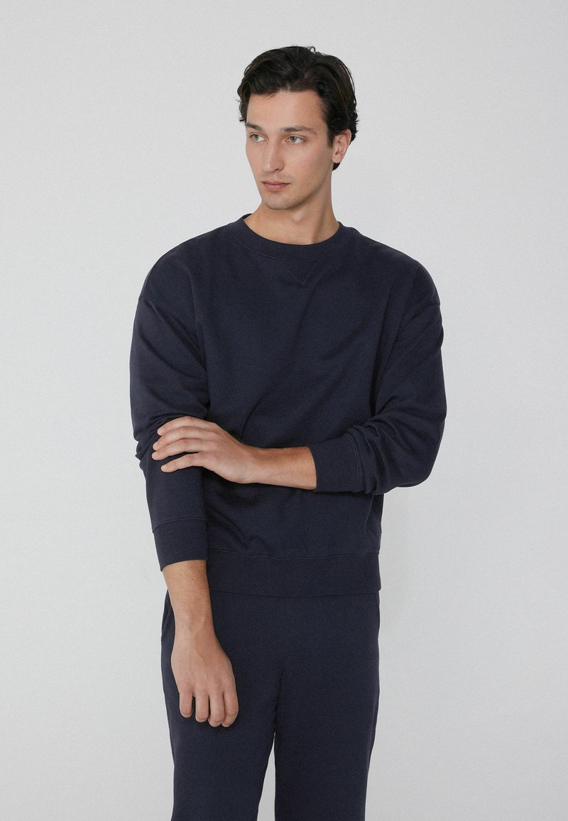 Sudadera de cuello redondo en color navy con tejido texturizado, puños y dobladillo de canalé. Modelo vistiendo con pantalones a juego, brazos cruzados.