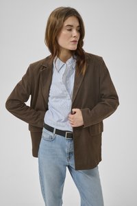 Vrouw met een bruin suède colbert over een blauw gestreept overhemd en lichtblauwe spijkerbroek, die met één hand het colbert rechtzet en opzij kijkt.