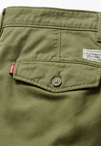 Olivgrön tyg med en slät textur, med en bakficka med knappstängning och en etikett sydd på midjebandet.