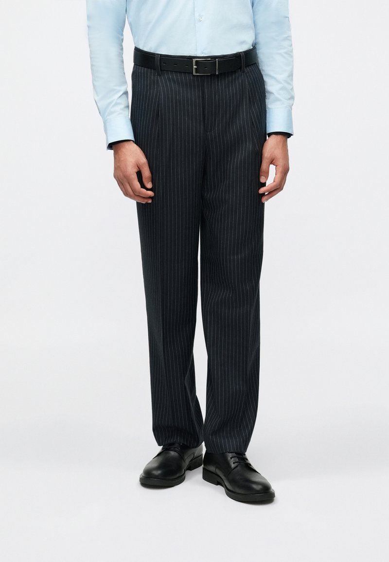 Calças escuras risca de giz feitas de tecido, com um design de perna reta, passadores de cinto e um cinto de couro preto, combinadas com sapatos pretos.