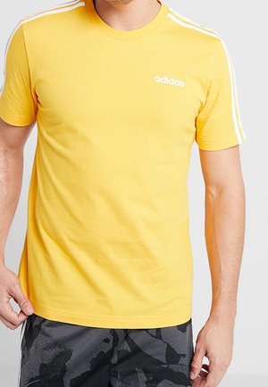 Man met een geel Adidas T-shirt met witte strepen op de mouwen en zwarte korte broek met patroon, tegen een effen achtergrond.