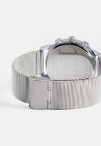 Silver stainless steel watch with a mesh strap, featuring a round case and a polished finish. The back displays manufacturer details.