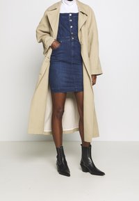 Beige lång kappa över en mörk denimklänning med knappar och sidofickor, kombinerad med svarta ankelstövlar med spetsig tå och klossklack.