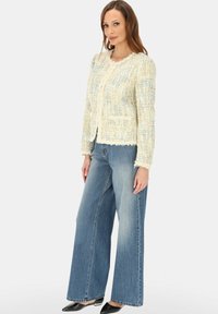 Kobieta stojąca, ubrana w kremową, teksturowaną kurtkę z frędzlami, szerokie niebieskie dżinsy i czarne buty na niskim obcasie na białym tle.