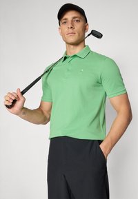 Grön polo med krage och tre knappar, matchad med svarta byxor. Håller en golfklubba; minimalistisch logotyp på skjortan.