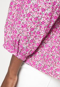 Manche de blouse florale rose vif avec poignet élastique et bouton, portée avec un pantalon blanc sur un bras à peau foncée, sur un fond uni.