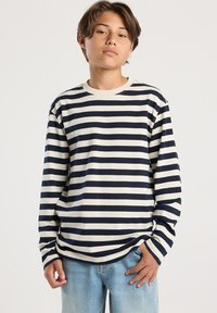 Teenager mit hellbraunem Haar, der ein schwarz-weiß gestreiftes Langarmshirt und hellblaue Jeans trägt, steht vor einem einfarbigen Hintergrund.