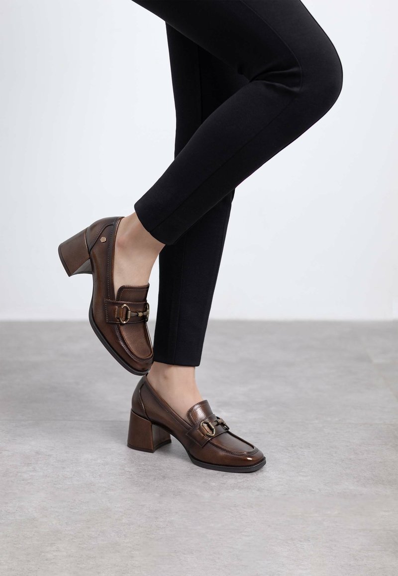 Mocasines de cuero marrón con tacón, punta cuadrada, hebilla metálica decorativa y un tacón grueso, combinados con pantalones negros ajustados.