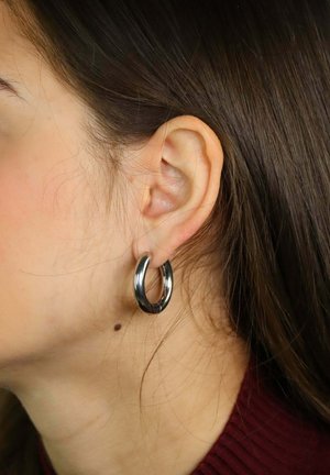 Nahaufnahme eines Ohrs einer Frau mit dunklem Haar, die einen dicken silbernen Creolenohrring trägt, sowie einem kleinen Muttermal an der Kieferlinie.