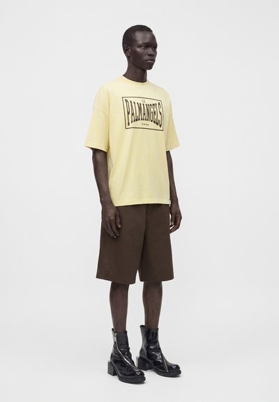 Άνδρας που φοράει άνετο ανοιχτό κίτρινο t-shirt με κείμενο "Palm Angels", καφέ σορτς μέχρι το γόνατο και μαύρα λαμπερά μπότες με τακούνι, στέκεται σε λευκό φόντο.