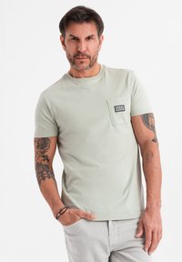 Camiseta de manga corta verde claro con un bolsillo en el pecho, textura suave y una pequeña etiqueta en el bolsillo. Presenta tatuajes visibles en los brazos.