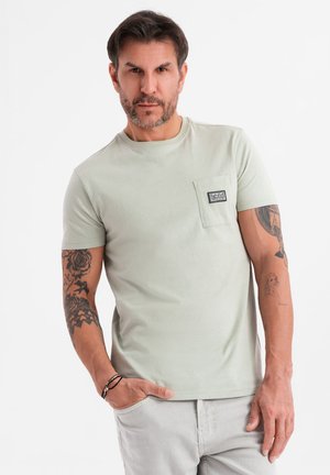 Camiseta de manga corta verde claro con un bolsillo en el pecho, textura suave y una pequeña etiqueta en el bolsillo. Presenta tatuajes visibles en los brazos.