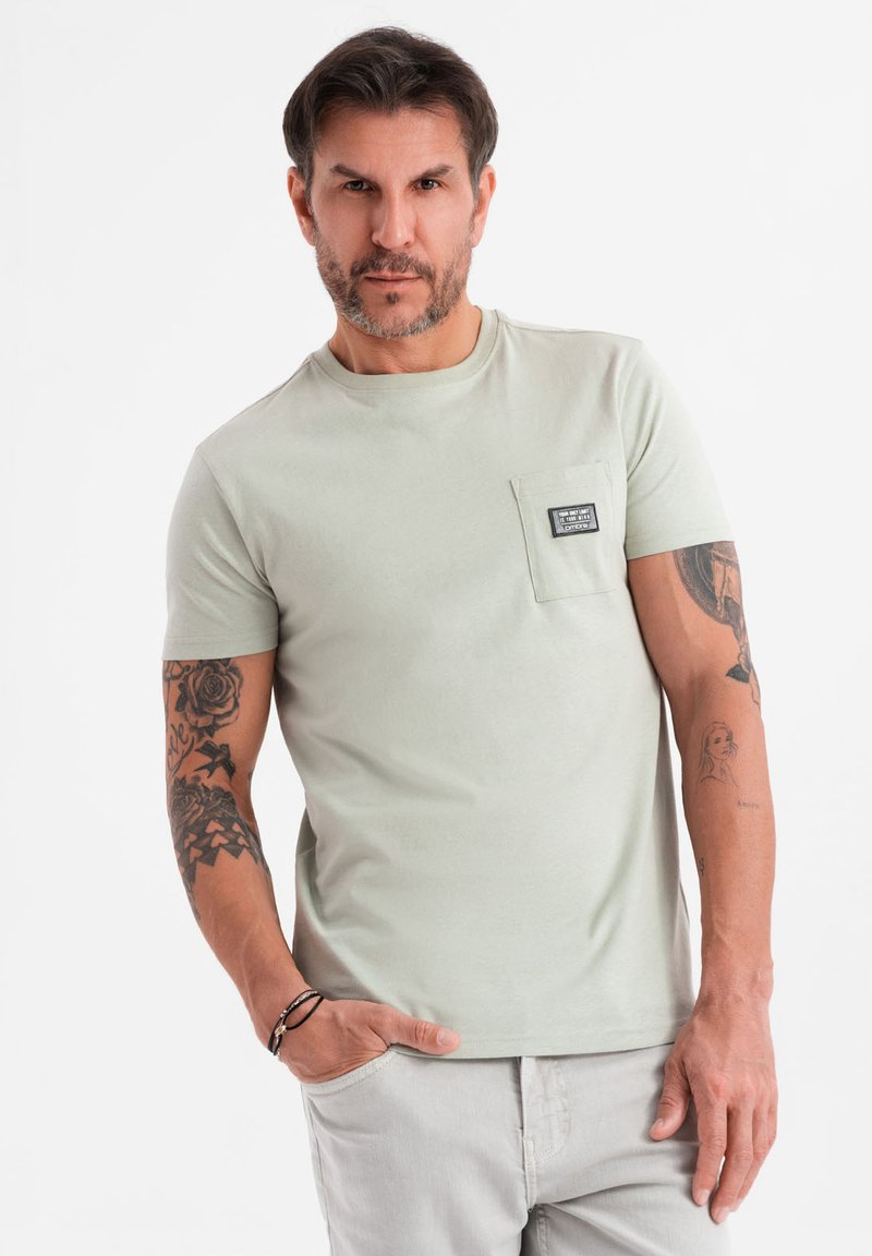 Lysegrøn kortærmet T-shirt med en brystlomme, blød tekstur og et lille mærke på lommen. Viser synlige tatoveringer på armene.