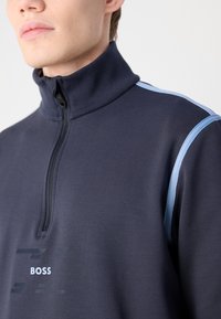 Il pullover navy a mezza zip presenta un col alto, strisce laterali azzurro chiaro e un logo stampato "BOSS" sul davanti. Tessuto morbido e strutturato.