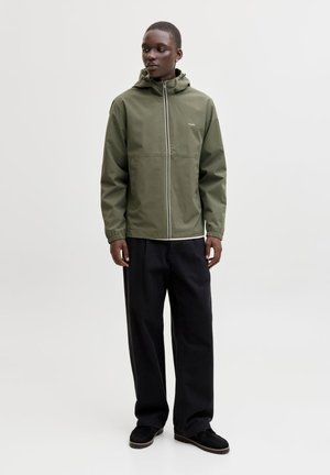 Junger Mann trägt eine olivgrüne Kapuzenjacke, schwarze weite Hosen und schwarze Schuhe, steht vor einem schlichten weißen Hintergrund.