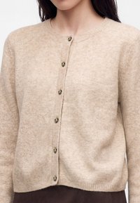 Beige strikket cardigan med knapper og rund halsudskæring, båret over en mørk underdel, viser en persons overkrop og nakke.