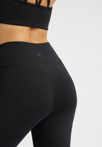 Leggings neri a vita alta realizzati in tessuto elasticizzato, caratterizzati da una texture liscia e un dettaglio con logo sulla cintura, che esaltano la forma.