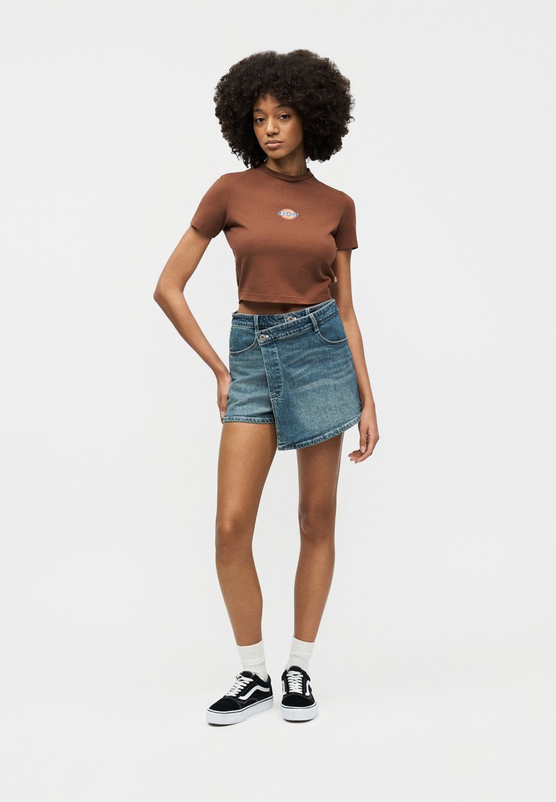 Donna con capelli ricci che indossa una t-shirt marrone corta, una gonna di jeans asimmetrica, calze bianche e sneakers nere, in piedi davanti a uno sfondo semplice.