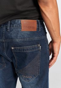 Donkerblauwe spijkerbroek van denim met een leren patch, complexe stiksels en ritszakken aan de achterkant met een gestructureerd patroon en logo.