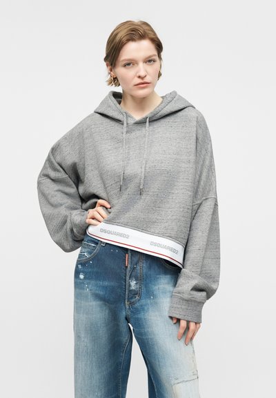 Νεαρή γυναίκα με κοντά μαλλιά, που φοράει μια γκρι φαρδιά μπλούζα με κουκούλα με τη λέξη "DSQUARED2" στο τελείωμα και ξεθωριασμένο μπλε τζιν με φθορές.