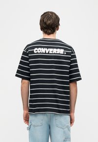 Άτομο που φοράει κοντομάνικη μπλούζα με μαύρες και λευκές ρίγες και το κείμενο "CONVERSE" στην πλάτη, συνδυασμένη με ανοιχτόχρωμο τζιν παντελόνι.