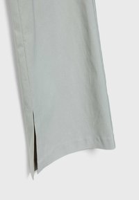 Gros plan sur une jambe de pantalon en tissu gris clair avec une petite fente latérale près de l'ourlet sur un fond blanc uni.