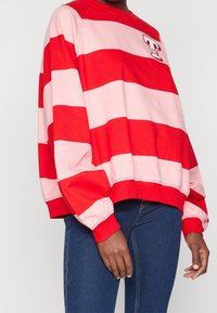 Felpa a righe rosse e rosa con collo a giro e maniche a sbuffo. Presenta un logo ricamato sul petto. Indossata con jeans blu scuro.