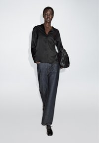 Czarna satynowa koszula z guzikami i luźne ciemnogranatowe spodnie. Model trzyma czarną skórzaną torbę. Obuwie to błyszczące czarne buty.