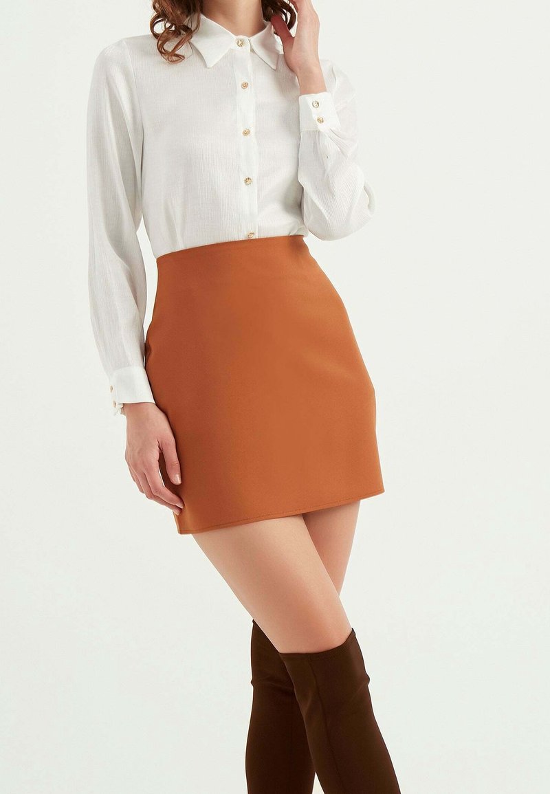 Mujer con blusa blanca de manga larga abotonada, minifalda color óxido y botas altas marrón oscuro, posando contra un fondo liso.