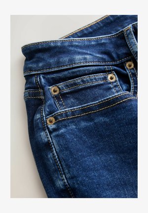 Nærbillede af blå denimjeans med forlomme, nitter, syninger og linning på en hvid baggrund.