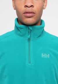 Mladenič oblečen v teal flis pulover s četrtno zadrgo in majhnim logotipom "HH" na levem prsnem delu.