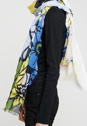 Personne portant un haut à manches longues noir et une écharpe florale colorée aux tons bleu, vert, jaune et blanc, drapée autour du cou.