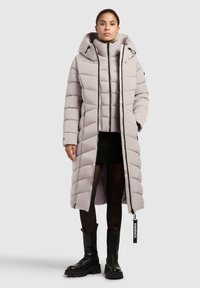 Svetlošedý dlhý vypchávaný kabát s kapucňou, ktorý má vertikálny prešívaný dizajn, zips a predné rozparky. Nosí sa cez priliehavý top a sukňu.