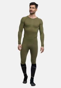 Homme portant un haut à manches longues moulant vert olive et un legging assorti, accompagnés de chaussettes hautes noires et grises, se tenant sur un fond blanc.