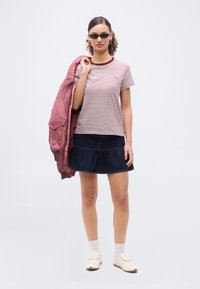 Γυναίκα που φοράει ριγέ μπλουζάκι, σκούρα τζιν φούστα, λευκά παπούτσια, κρατώντας ροζ μπουφάν στον ώμο και μαύρα γυαλιά ηλίου μπροστά σε απλό φόντο.