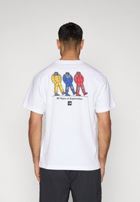 Λευκό βαμβακερό μπλουζάκι με γραφικό τριών φιγούρων σε κίτρινα, μπλε και κόκκινα μπουφάν, με το κείμενο «60 Χρόνια Εξερεύνησης» από κάτω.