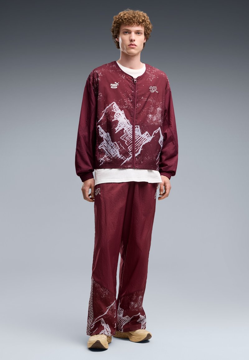 Jeune homme aux cheveux bouclés porte un survêtement bordeaux avec des motifs de montagnes blancs, une chemise blanche en dessous et des baskets beiges, debout sur un fond gris.